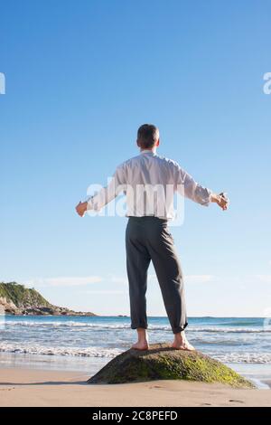 Homme d'affaires mature qui profite du soleil tout en se tenant sur un rocher sur une plage au lever du soleil Banque D'Images