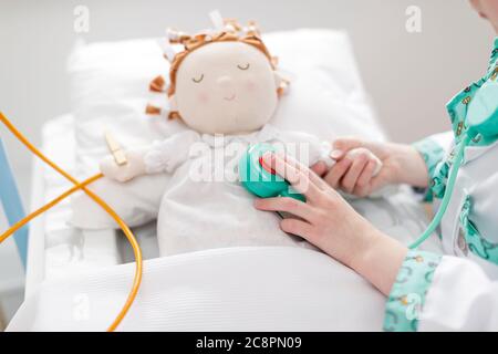 Jeune fille vêtue comme médecin utilisant le stéthoscope jouet sur la poupée dans le lit d'hôpital de make-Believe Banque D'Images