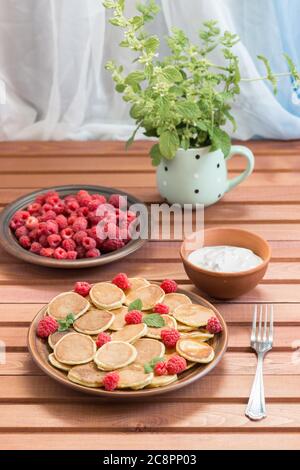 Céréales pancakes et framboises fraîches mûres rouges dans une assiette brune. Un bouquet de Melissa dans une tasse sur une table en bois sombre près de la fenêtre. Un été délicieux Banque D'Images