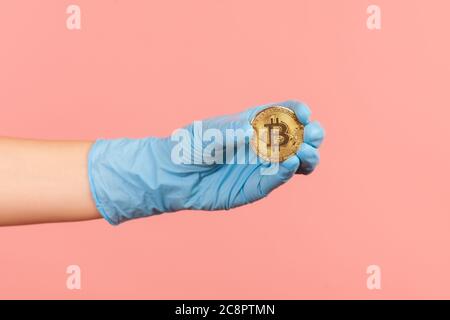 Vue latérale du profil gros plan de la main humaine dans des gants chirurgicaux bleus tenant et montrant le symbole bitcoin en main. Vue intérieure, prise en studio, isolée sur le dos rose Banque D'Images