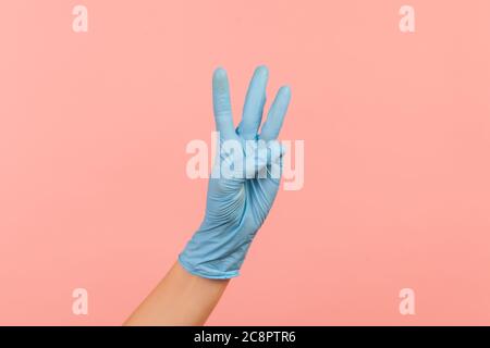 Vue latérale du profil gros plan de la main humaine avec des gants chirurgicaux bleus, portant le numéro 3 trois avec les mains. Vue en studio, isolée sur fond rose. Banque D'Images