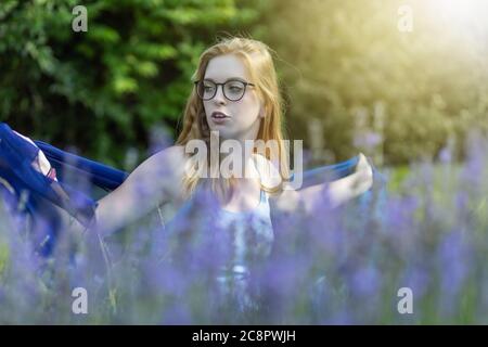 Une fille à cheveux longs avec des lunettes joue avec un foulard bleu derrière la lavande dans le jardin Banque D'Images