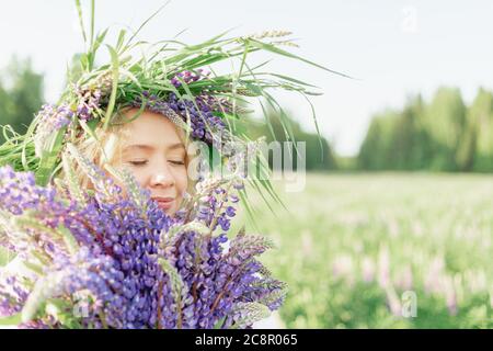 Une fille blonde dans une couronne de lupin de fleur avec un bouquet de lupins sourit doucement et inonde le parfum des fleurs dans un pré de fleur sur un Soleil Banque D'Images