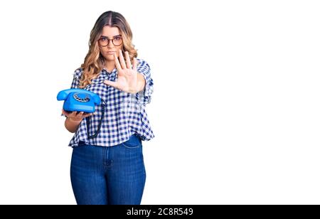 Jeune femme caucasienne tenant téléphone vintage avec main ouverte faisant signe d'arrêt avec expression sérieuse et confiante, geste de défense Banque D'Images