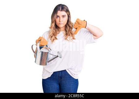 Jeune femme caucasienne portant des gants portant l'arrosoir peut avec visage en colère, signe négatif montrant le désarroi avec les pouces vers le bas, concept de rejet Banque D'Images