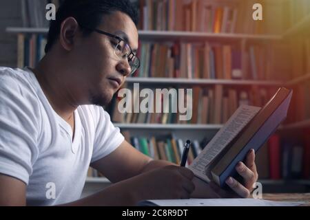 Jeune homme asiatique étudiant l'apprentissage en bibliothèque, concept de préparation à l'examen. Étudiant d'université de sexe masculin faisant de la recherche et prenant des notes dans son livre Banque D'Images