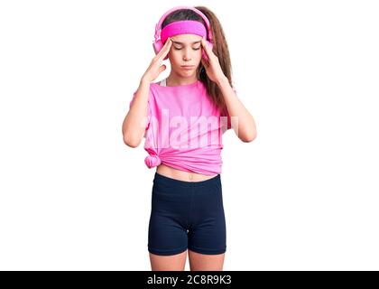 Mignon hispanique enfant fille portant des vêtements de gym et utilisant des écouteurs avec main sur la tête pour la douleur dans la tête parce que le stress. Souffrant de migraine. Banque D'Images