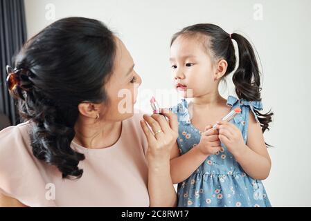 Curieuse adorable petite fille vietnamienne regardant sa mère appliquant un rouge à lèvres rose clair quand se préparer le matin Banque D'Images