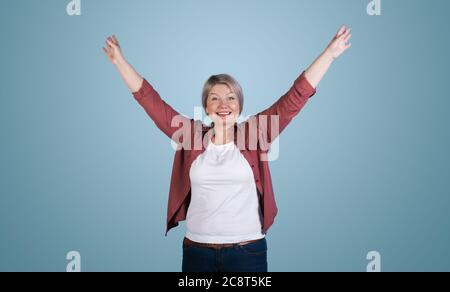 Femme blonde senior posant les mains vers le haut et souriant édenieusement sur un mur bleu de studio Banque D'Images