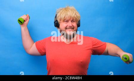 Drôle de gras homme freaky dans une perruque et un t-shirt rose avec des haltères faisant des exercices sur un fond bleu. Banque D'Images