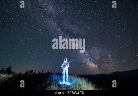 Un spaceman lumineux dans un costume d'espace blanc debout au milieu d'une prairie de montagne tenant une guitare, ciel nocturne incroyable plein d'étoiles sur fond. Concept de musique, de cosmonautics et de voyages spatiaux. Banque D'Images