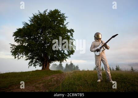 Astronaute guitariste en costume spatial et casque avec instrument de musique jouant de la guitare, debout dans un pré à flanc de colline avec un magnifique arbre et le ciel sur fond. Concept de musique, de cosmonautics et de nature. Banque D'Images