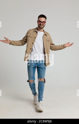 jeune homme décontracté non rasé en veste portant des lunettes de soleil, ouvrant les bras et accueillant, marchant et posant sur fond gris Banque D'Images