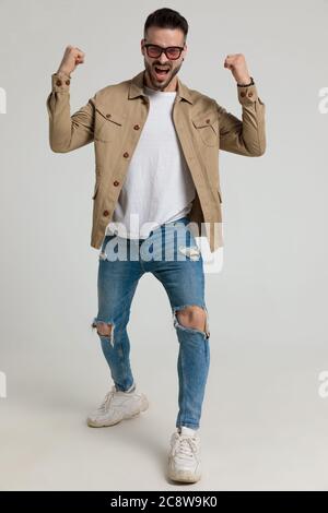 un jeune homme enthousiaste en veste portant des lunettes de soleil, tenant les poings dans l'air et criant, célébrant la victoire sur fond gris Banque D'Images