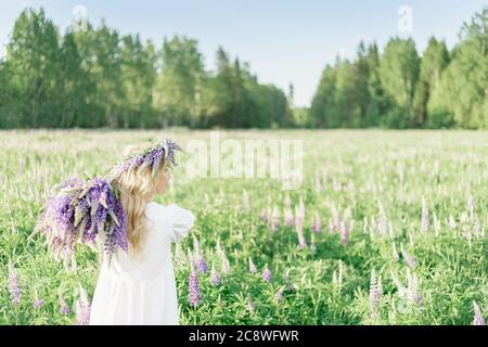 Une fille avec un bouquet de fleurs sur son épaule dans une robe blanche et une couronne de fleurs marche en arrière le long du champ de fleurs, dans le champ de fleurs de Banque D'Images