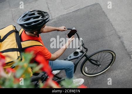 Vélo de coursier dans un casque de protection avec sac à dos de livraison se trouve sur le vélo et regarde le téléphone et utilise la carte en ligne sur la rue Banque D'Images