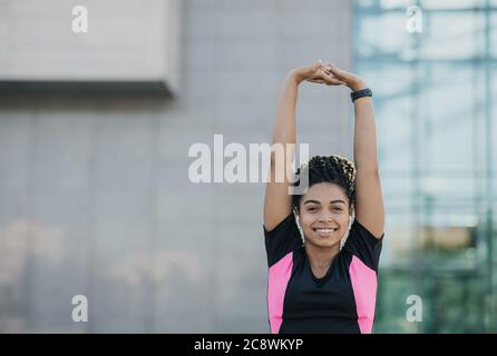 Souriant afro-américaine fille dans les vêtements de sport avec casque sans fil et tracker d'activité faisant étirage pour les mains Banque D'Images