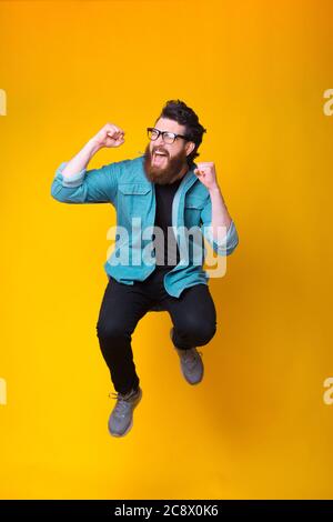 Happy Bearded hipster saute dans un studio en faisant un geste gagnant tout en criant sur fond jaune. Banque D'Images