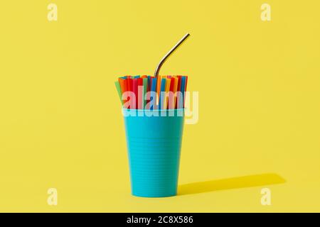 une paille réutilisable en acier inoxydable se distingue de quelques pailles jetables en plastique de différentes couleurs dans un vase bleu, sur un bac jaune Banque D'Images