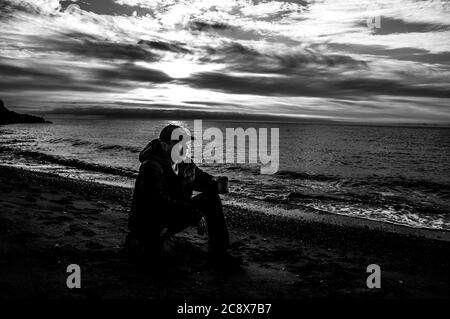 Tasse de thé le matin. Homme assis sur un rocher et buvant du thé au lever du soleil sur une plage de mer. Noir et blanc, contraste élevé. Banque D'Images