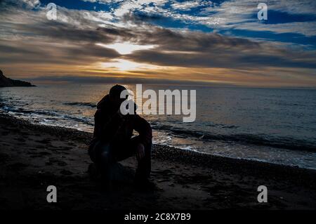 Tasse de thé le matin. Homme assis sur un rocher et buvant du thé au lever du soleil sur une plage de mer. Banque D'Images