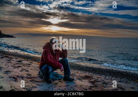 Tasse de thé le matin. Homme assis sur un rocher et buvant du thé au lever du soleil sur une plage de mer. Banque D'Images