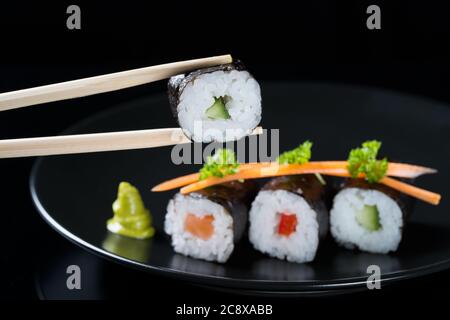 Bâtonnets tenant le rouleau avec le concombre sur fond de rouleaux frais avec le saumon, le concombre et le poivre sur une assiette, servis avec des tranches de carottes et du persil, Banque D'Images