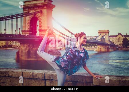 Jeune femme à tête rouge faisant du yoga au Danube à la lumière de la ville, pose de cygne, Budapest, Hongrie Banque D'Images