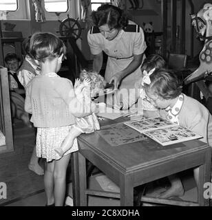 années 1950, historique, une infirmière de navire en uniforme avec de jeunes enfants dans la pépinière d'un paquebot. Une jeune fille lit un livre d'images, tandis qu'une autre tient une poupée jouet. Banque D'Images