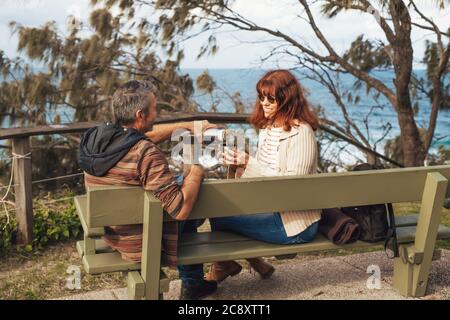 Joyeux couple d'âge moyen amoureux assis sur le banc dans le parc, sourire et boire du thé ou du café de thermos. Banque D'Images