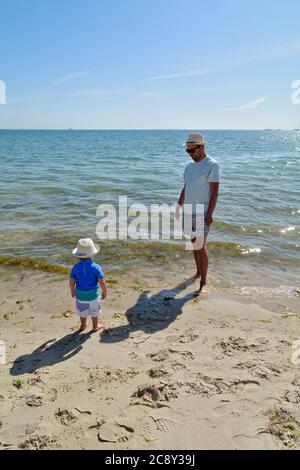 Un père et un fils tout-petits debout sur le bord de l'eau à la plage de Studland Bay, un jour d'été chaud, Dorset Angleterre Royaume-Uni Banque D'Images