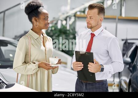 un vendeur caucasien amical parle avec une cliente noire, un agent commercial professionnel parle des avantages et des caractéristiques de la voiture, une femme écoute t Banque D'Images