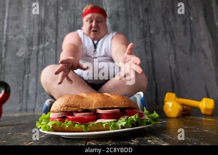 obèse jeune homme caucasien empiète sur un gros sandwich, l'homme gras ne peut pas adhérer à un mode de vie sain et à une alimentation Banque D'Images