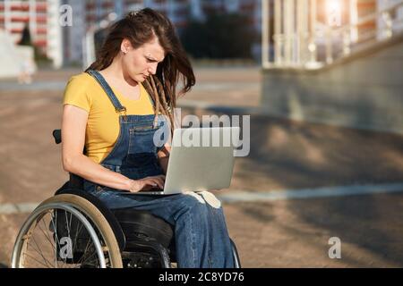 élégante fille handicapée dans des vêtements décontractés concentré sur le travail avec ordinateur portable dans la rue, gros plan vue latérale photo, espace de copie, arrière-plan flou. Banque D'Images