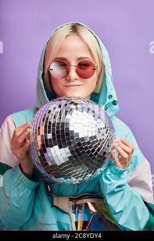 Portrait de belle belle blonde hipster femme dans ses années 20 posant avec la discothèque brillante, vêtu de menthe tendance et de couleur rose veste de pluie. Banque D'Images