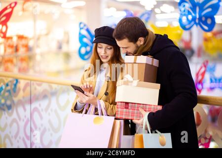 une femme caucasienne à poil dur avec des sacs d'achats montre quelque chose dans un smartphone à un homme barbu tenant des cadeaux, discuter de shopping à l'intérieur du centre commercial Banque D'Images