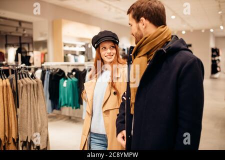 femme à poil rouge et homme à barbe caucasien en manteaux marcher dans le hall de la boutique de vêtements ensemble, choisir une nouvelle tenue Banque D'Images