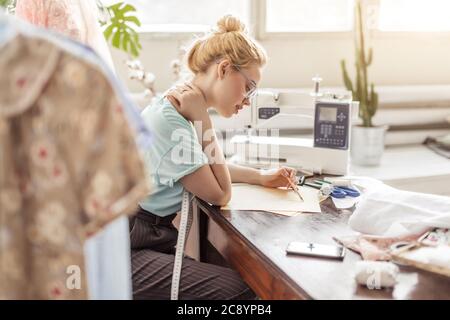 Créateur de mode sur mesure Belle de mesure se situe à dessin en milieu de travail ou de mettre par écrit les résultats après la mesure. Son studio est ensoleillée, pleine de colorf Banque D'Images