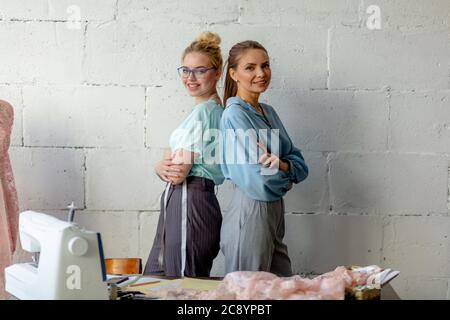 Portrait de deux jolies jeunes femmes couturières et à la mode dans des tons pastel, standing back to back and looking at camera dans bespoke clothi Banque D'Images