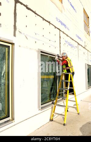 Un vitrier sur une échelle, appliquant du mastic d'étanchéité autour des bords des fenêtres nouvellement installées sur un chantier de construction. Banque D'Images