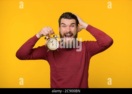 Photo d'un gars nerveux barbu, tenant une horloge et inquiet au sujet de la date limite, debout sur fond jaune Banque D'Images