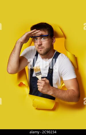 peintre caucasien travailleur en lunettes de sécurité et uniforme tenant la brosse de peinture et regardant vers l'avant à travers le trou dans le papier jaune. Banque D'Images