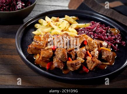 Assiette de goulash de bœuf allemand épicé au chou rouge et frites de pommes de terre sur une assiette noire servie à table en gros plan pour la publicité au menu Banque D'Images