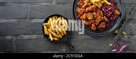 Bannière panoramique avec assiette de goulash de bœuf servie avec du chou rouge et une assiette de frites croustillantes sur fond de brique noire Banque D'Images