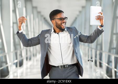 Homme d'affaires joyeux secouant les Fists tenant un smartphone et une tablette à l'aéroport Banque D'Images