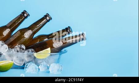 Bière fraîche le jour chaud. Flacons en verre foncé sur plateau en verre avec glace et citron vert sur fond bleu Banque D'Images