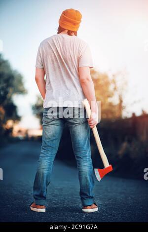 Un jeune homme avec un chapeau jaune tendance et un Jean bleu se tient avec confiance avec une grosse hache dans ses mains et regarde dans la distance. Survie. R Banque D'Images