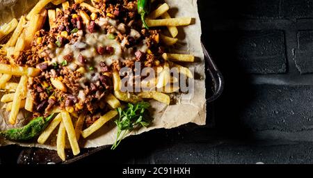 Frites de pommes de terre avec hache de bœuf épicée assaisonnée, piments, herbes et fromage servis sur du papier brun sur une casserole dans une vue de dessus sur un carrelage noir Banque D'Images