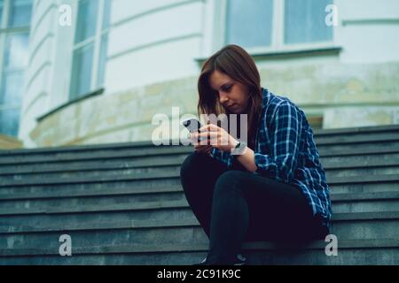 Une jeune femme qui fait des nouvelles dans un smartphone, assise dans un escalier de manoir. Femme adulte avec téléphone portable assise sur les marches du palais. Banque D'Images