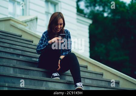 Une jeune femme qui fait des nouvelles dans un smartphone, assise dans un escalier de manoir. Femme adulte avec téléphone portable assise sur les marches du palais. Banque D'Images